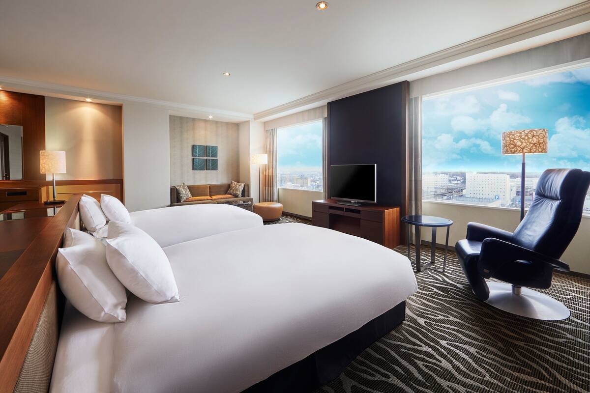 A hotel room with a large bed, a flat-screen TV, a seating area with a chair and table, and a city view through the window.