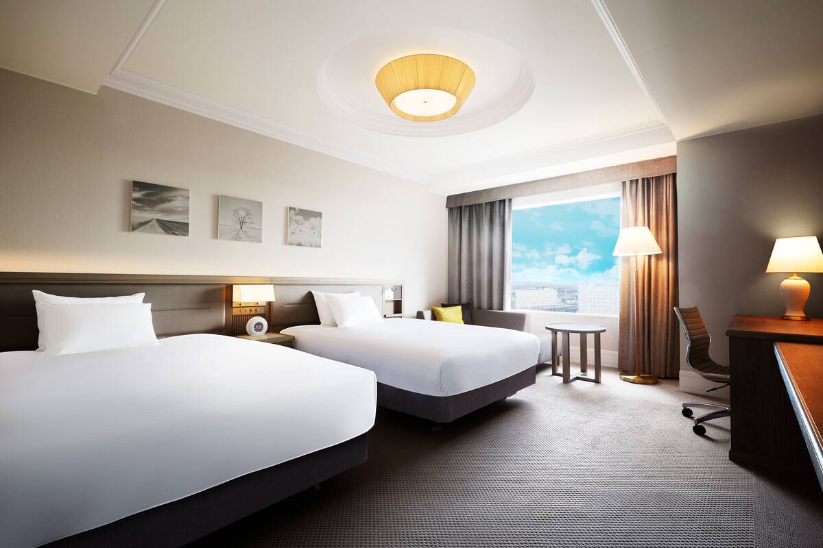 A hotel room with two beds, a desk, a chair, a small table, and a view of the sky through the window.