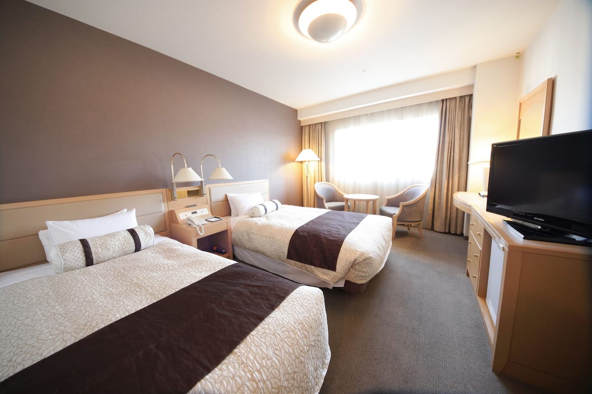 A hotel room with two beds, a desk, a television, and a window with curtains.