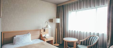 Habitación de hotel con cama, mesita de noche con lámpara, una silla y un ventanal con cortinas.
