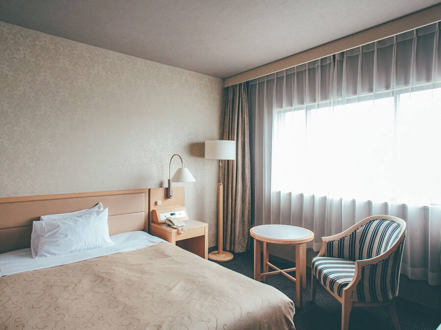 Habitación de hotel con cama, mesita de noche con lámpara, una silla y un ventanal con cortinas.