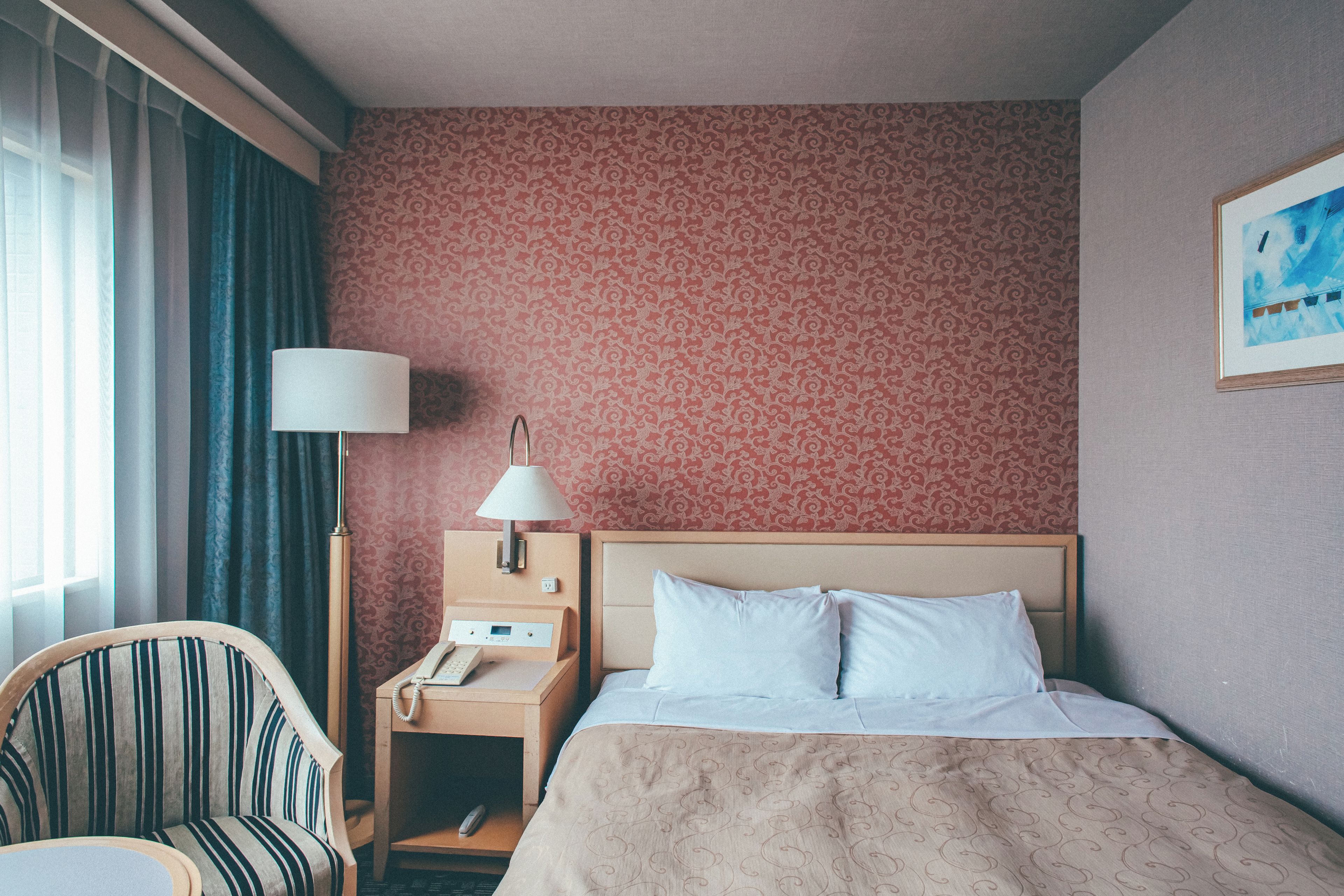 Una habitación de hotel con una cama, una silla, una mesita de noche con lámpara y un cuadro en la pared.