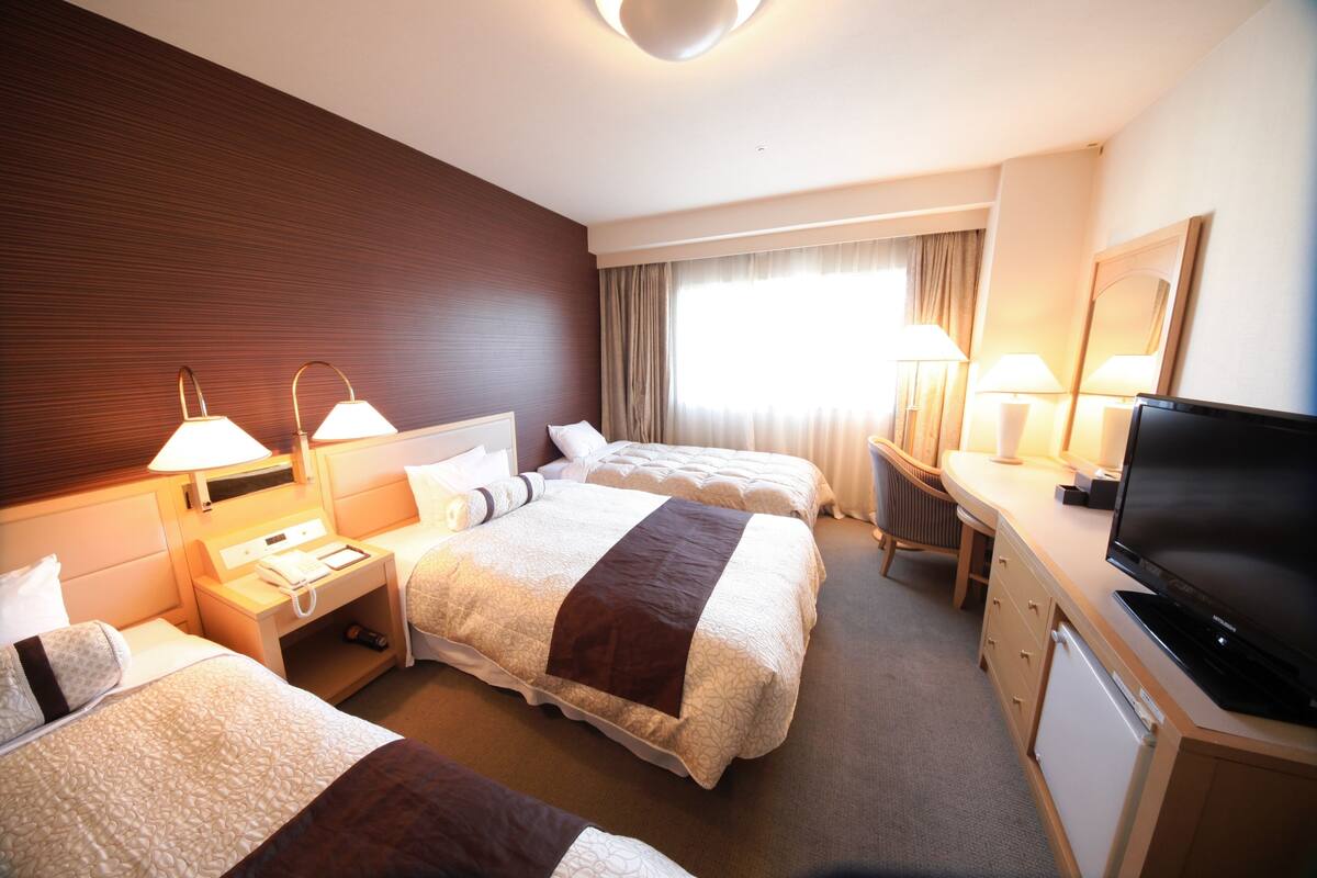A hotel room with two beds, a TV, a desk with a lamp, and a window with curtains.