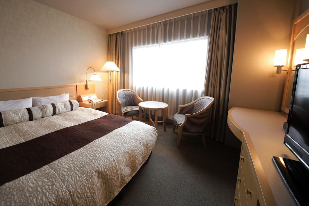 A hotel room with a large bed, two chairs, a small table, a desk with a TV, and a window with curtains.