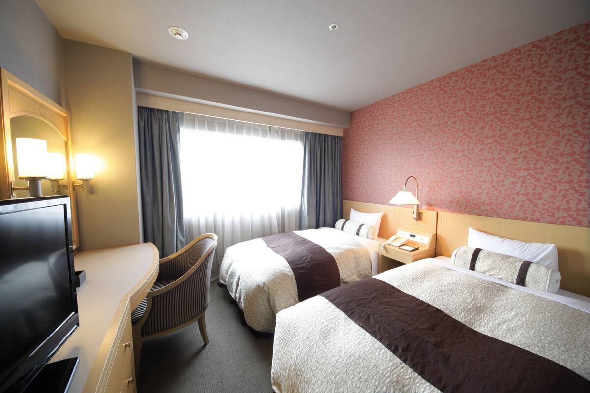 A hotel room with two beds, a desk with a TV, a chair, and a window with curtains.