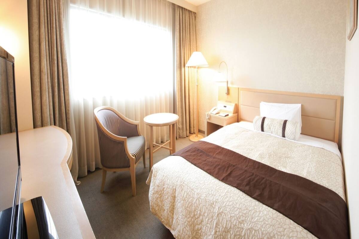 A hotel room with a bed, a chair, a small table, a lamp, and a telephone.