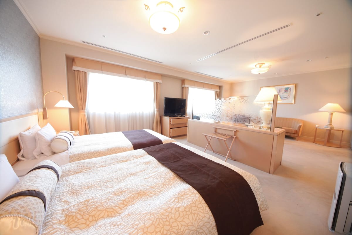 A hotel room with two beds, a desk, a TV, and a desk lamp.