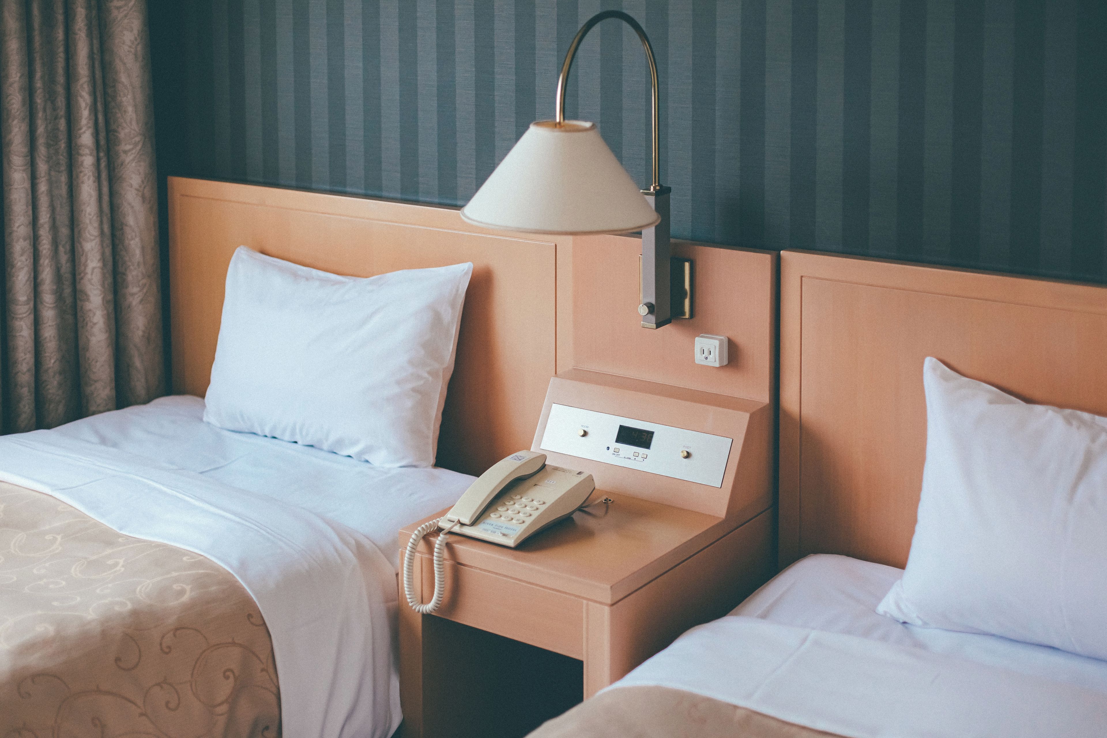 A hotel room with two beds, a bedside table with a lamp, a phone, and a control panel.