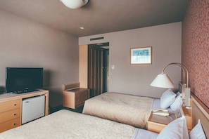 Ein Hotelzimmer mit zwei Betten, einem Schreibtisch mit Fernseher, einer Lampe und einem kleinen Kühlschrank.