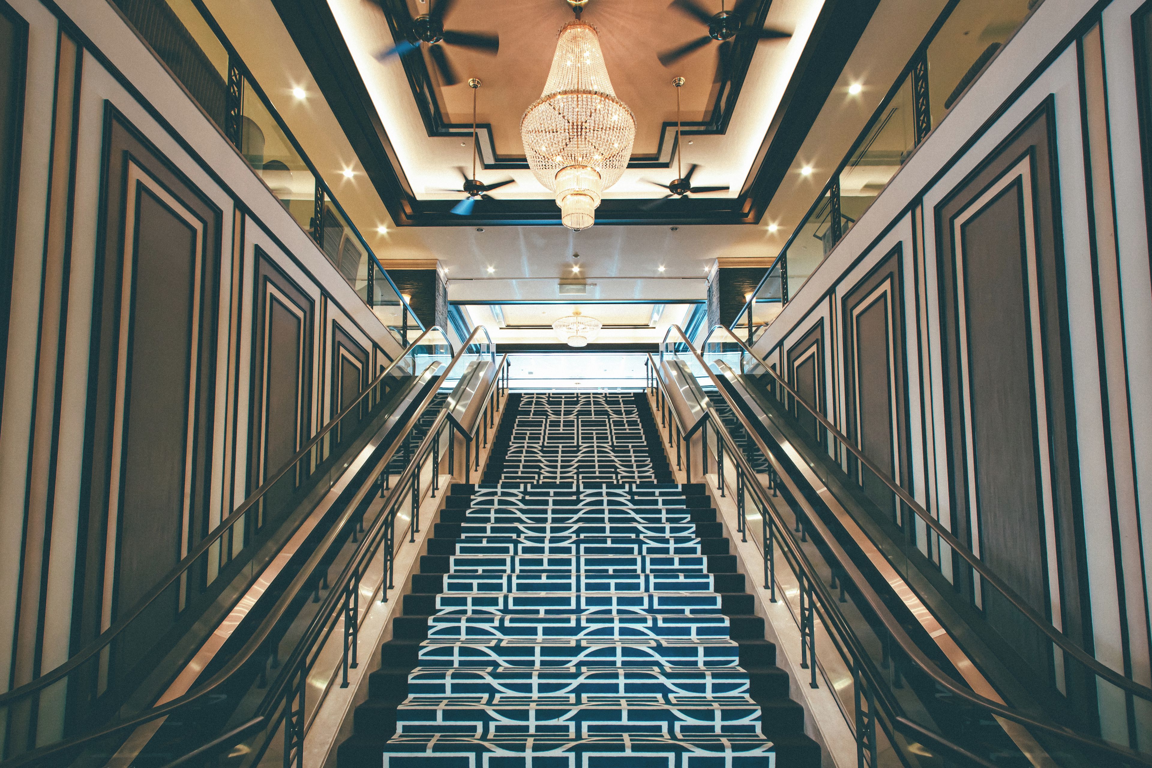 A grand staircase with a geometric patterned floor, flanked by glass railings and a central chandelier.