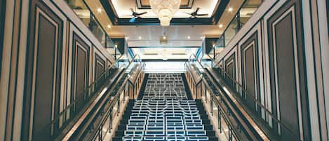 Una imponente escalera con piso de diseño geométrico, flanqueada por barandas de vidrio y un candelabro central.