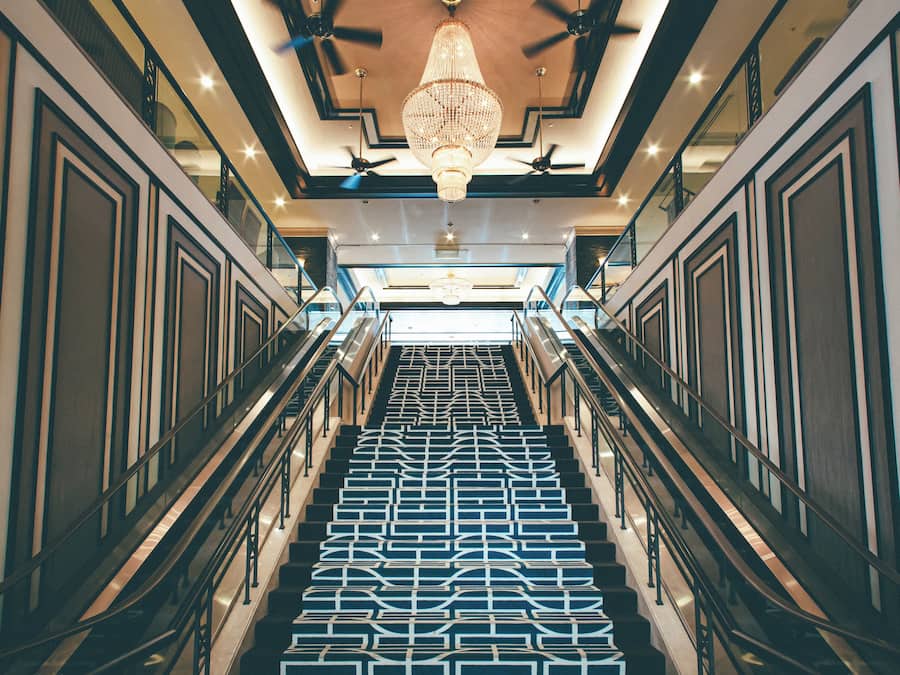 Una imponente escalera con piso de diseño geométrico, flanqueada por barandas de vidrio y un candelabro central.
