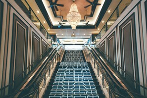 A grand staircase with a geometric patterned floor, flanked by glass railings and a central chandelier.