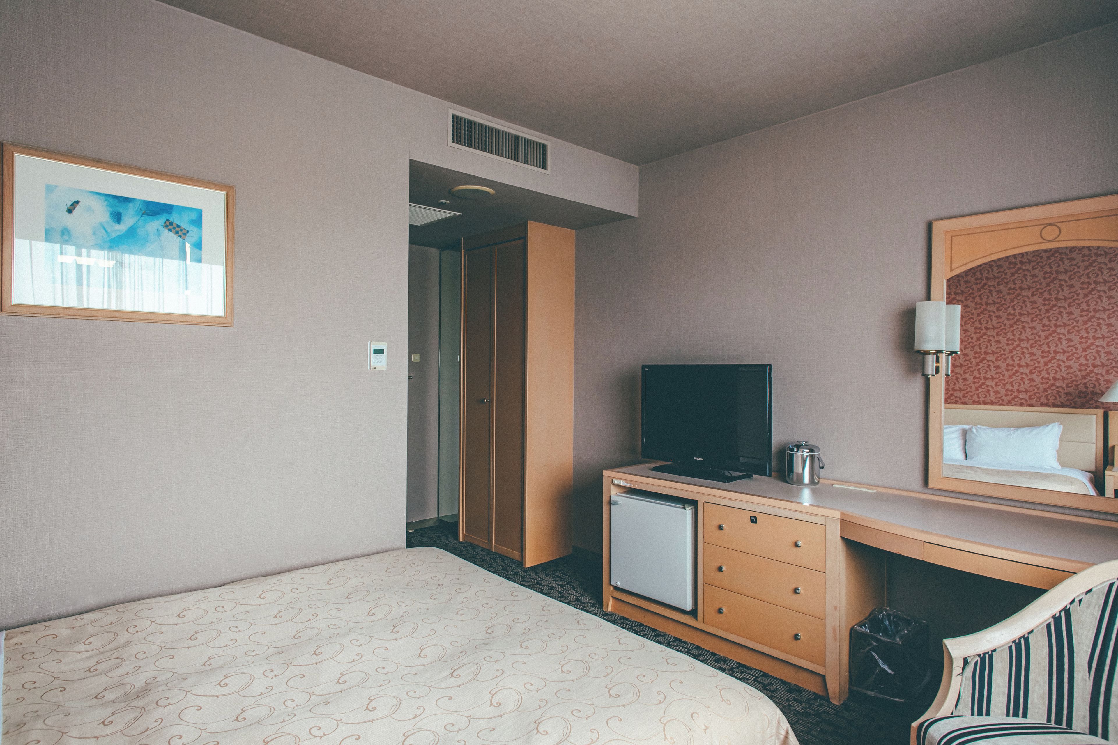 A hotel room with a bed, a desk with a television, a chair, a mirror, and a painting on the wall.