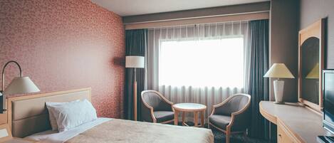 A hotel room with a bed, two chairs, a small table, a desk, a television, and a window with curtains.