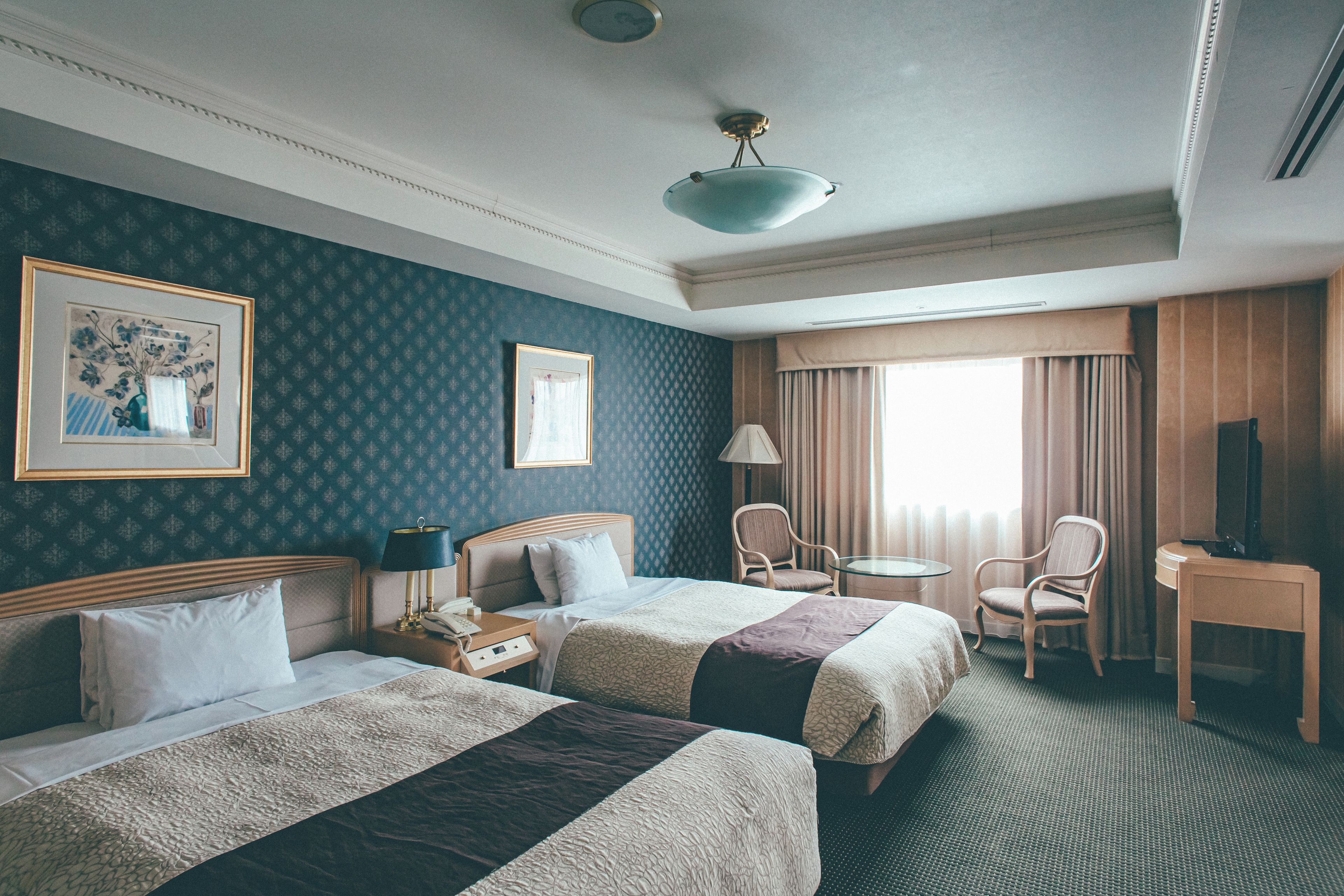A hotel room with two beds, a desk, a chair, a lamp, and a television.
