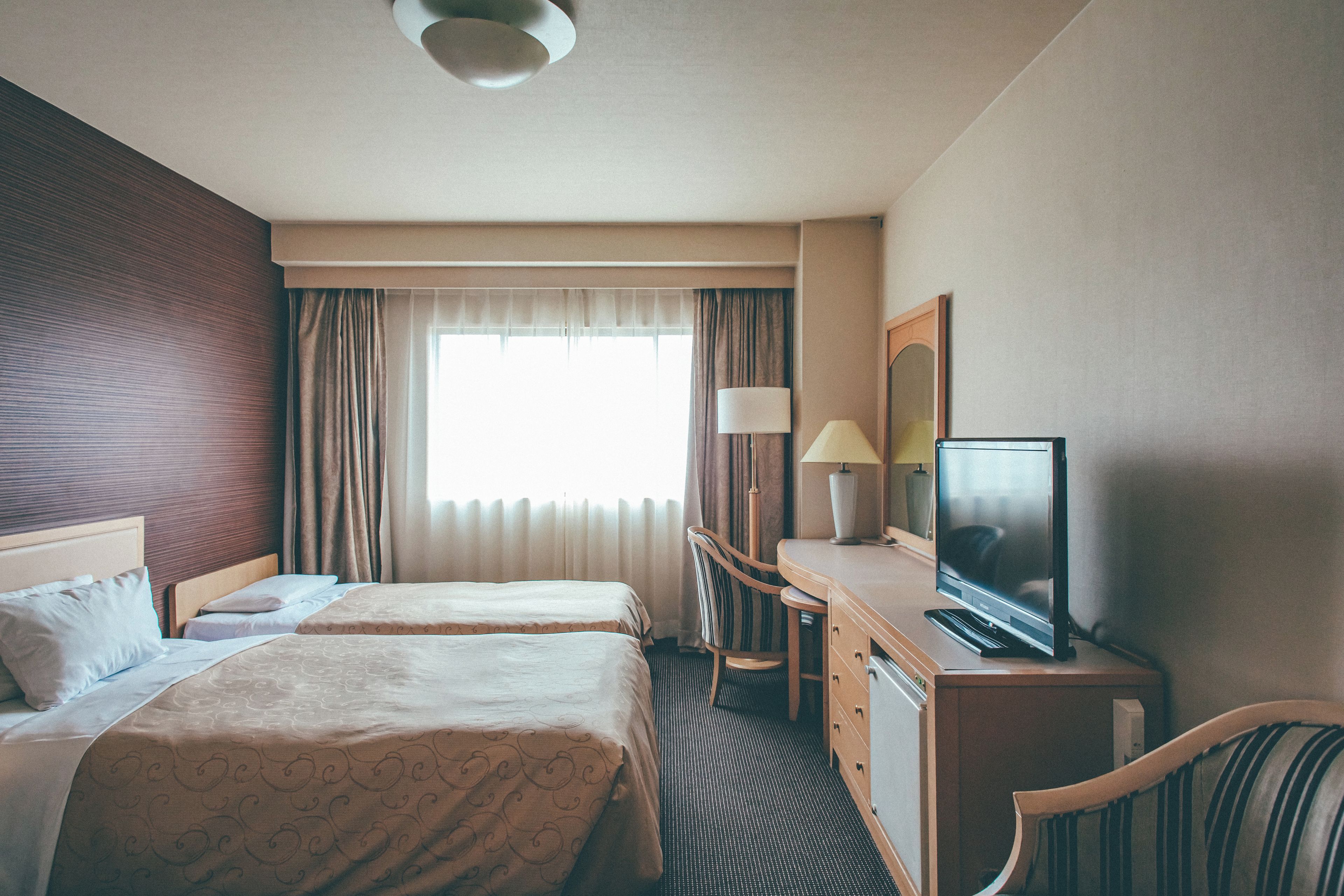 A hotel room with two beds, a desk with a television, a chair, and a window with curtains.