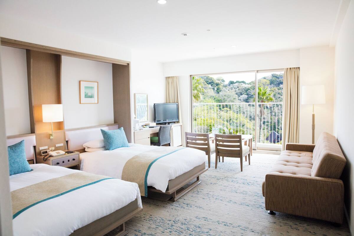A hotel room with two beds, a sofa, a TV, and a balcony with a view of greenery.