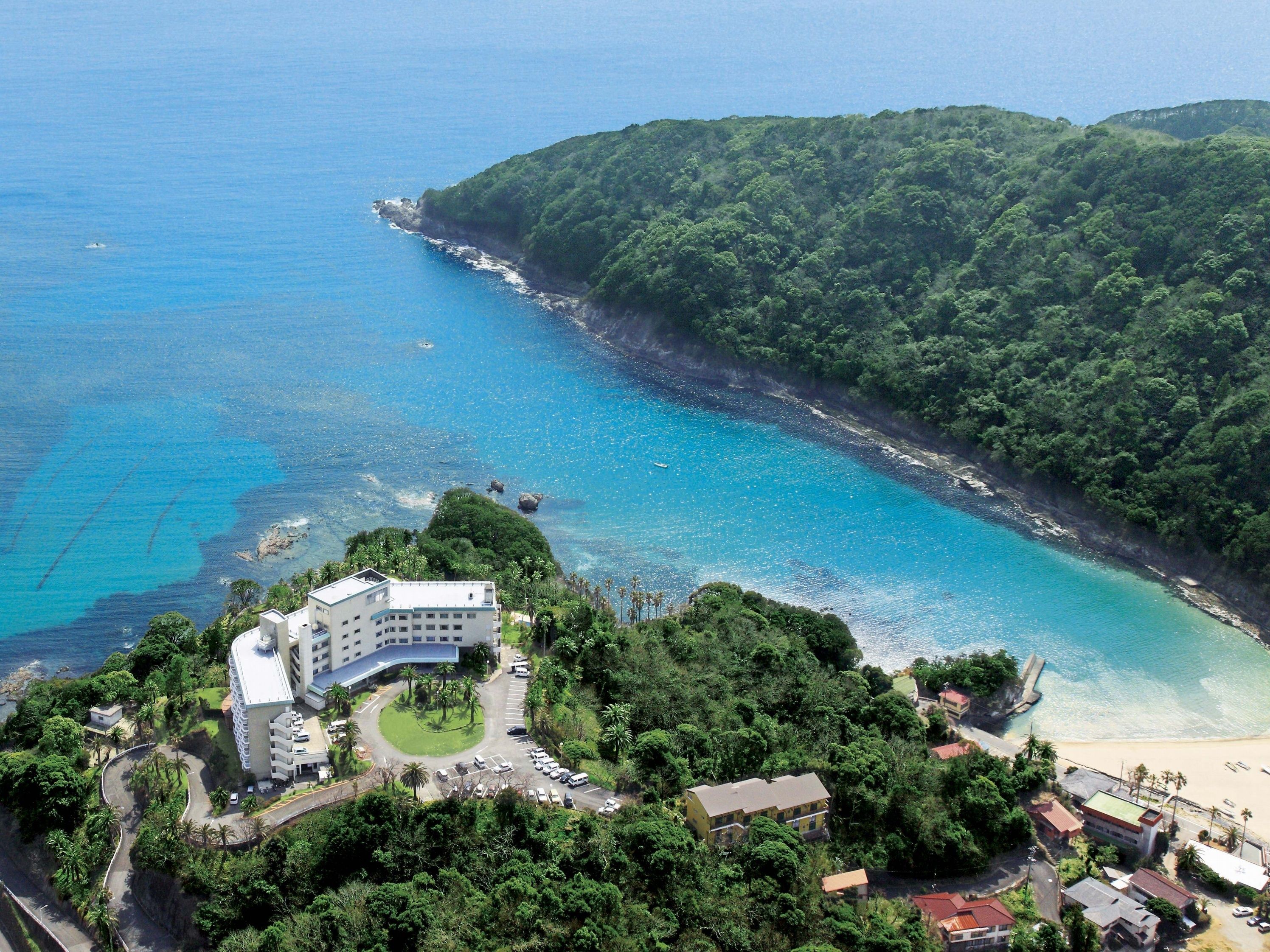 An aerial view of a coastal area with a hotel, lush greenery, and clear blue waters.