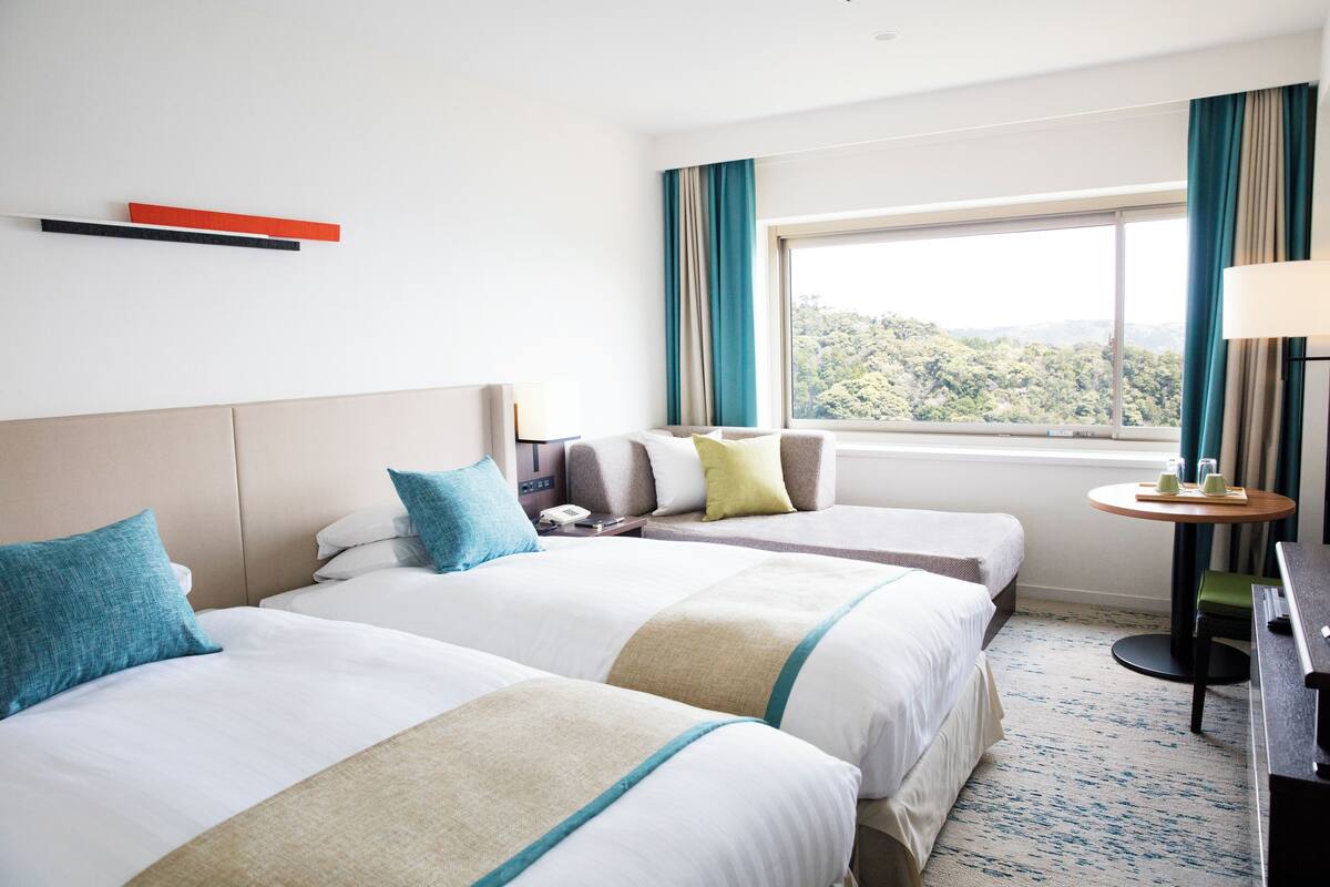 A hotel room with two beds, a desk, a chair, a window with a view of trees, and a table with a lamp.