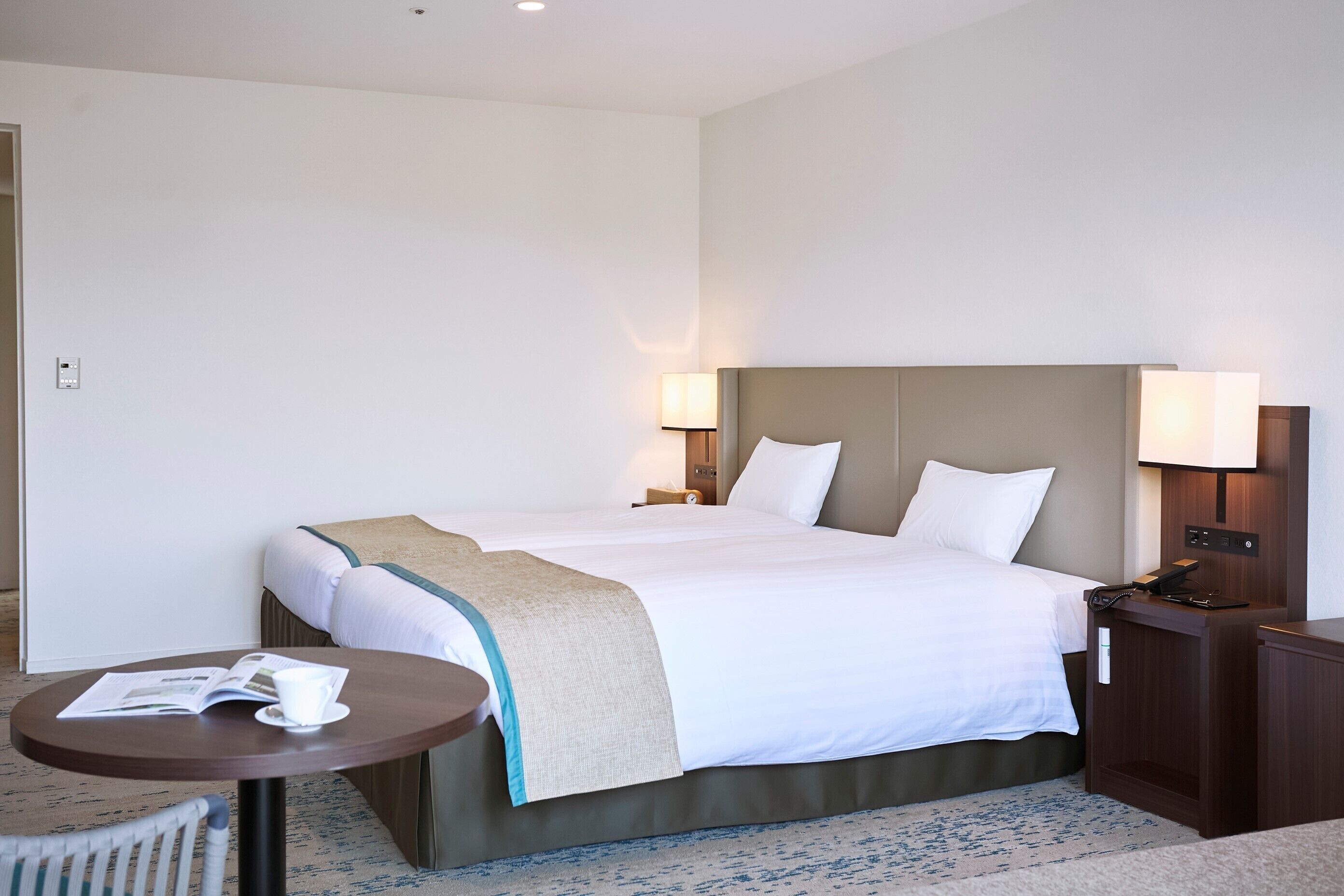 A hotel room with a large bed, bedside lamps, a small table with a magazine and a cup, and a wooden nightstand.