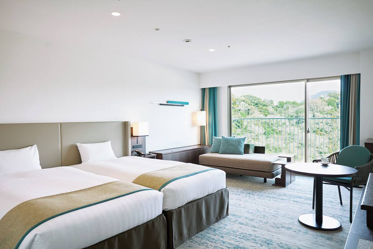 A hotel room with two beds, a sofa, a small table, and a balcony with a view of greenery.