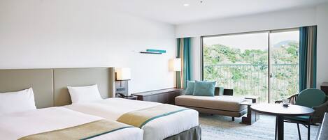 A hotel room with two beds, a sofa, a small table, and a balcony with a view of greenery.