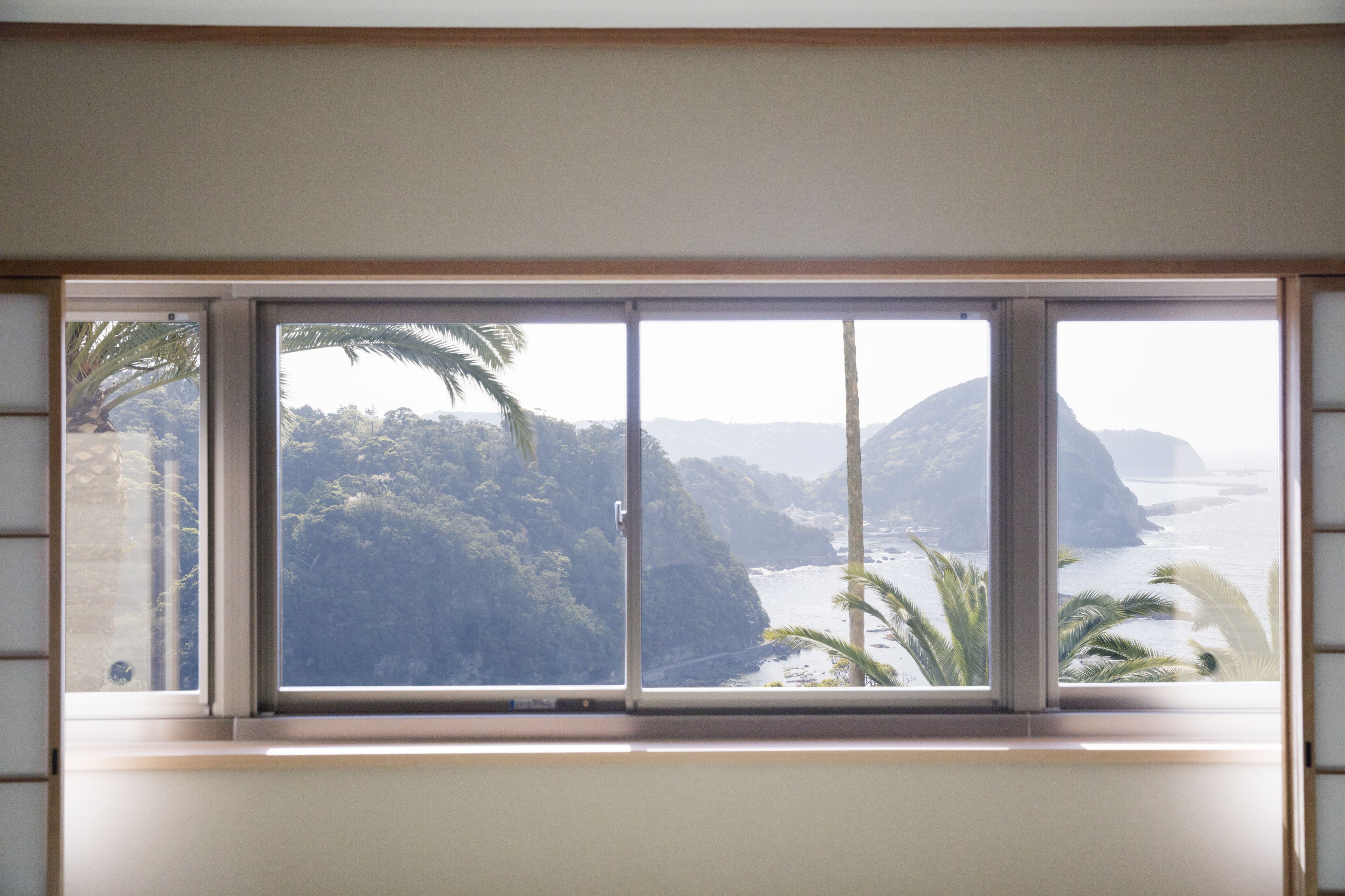 Ein Zimmer mit großem Fenster und Blick auf eine Küstenlandschaft mit Palmen und Klippen.