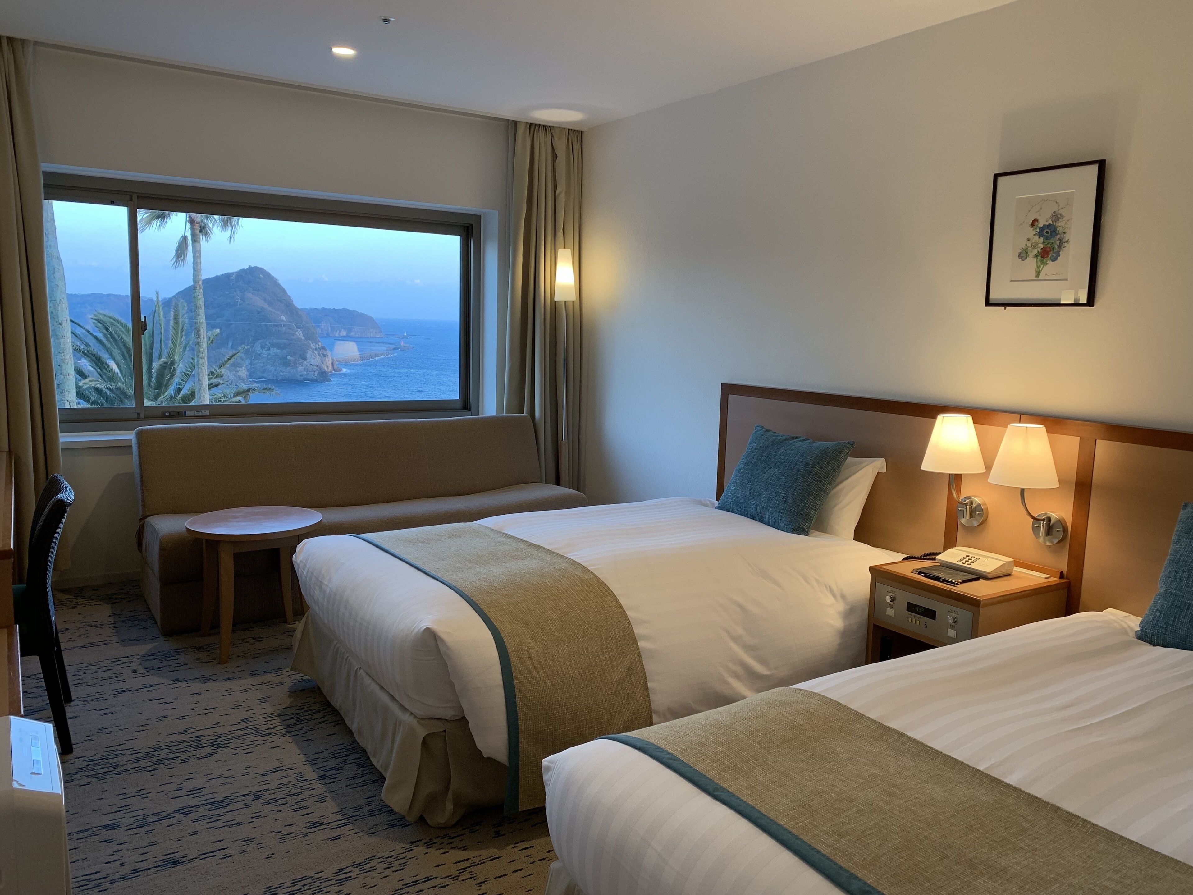 A hotel room with two beds, a seating area, a window with a view of the sea and a mountain, and a bedside table with a lamp.