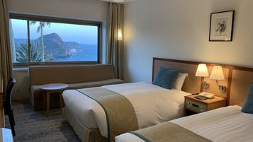 A hotel room with two beds, a seating area, a window with a view of the sea and a mountain, and a bedside table with a lamp.