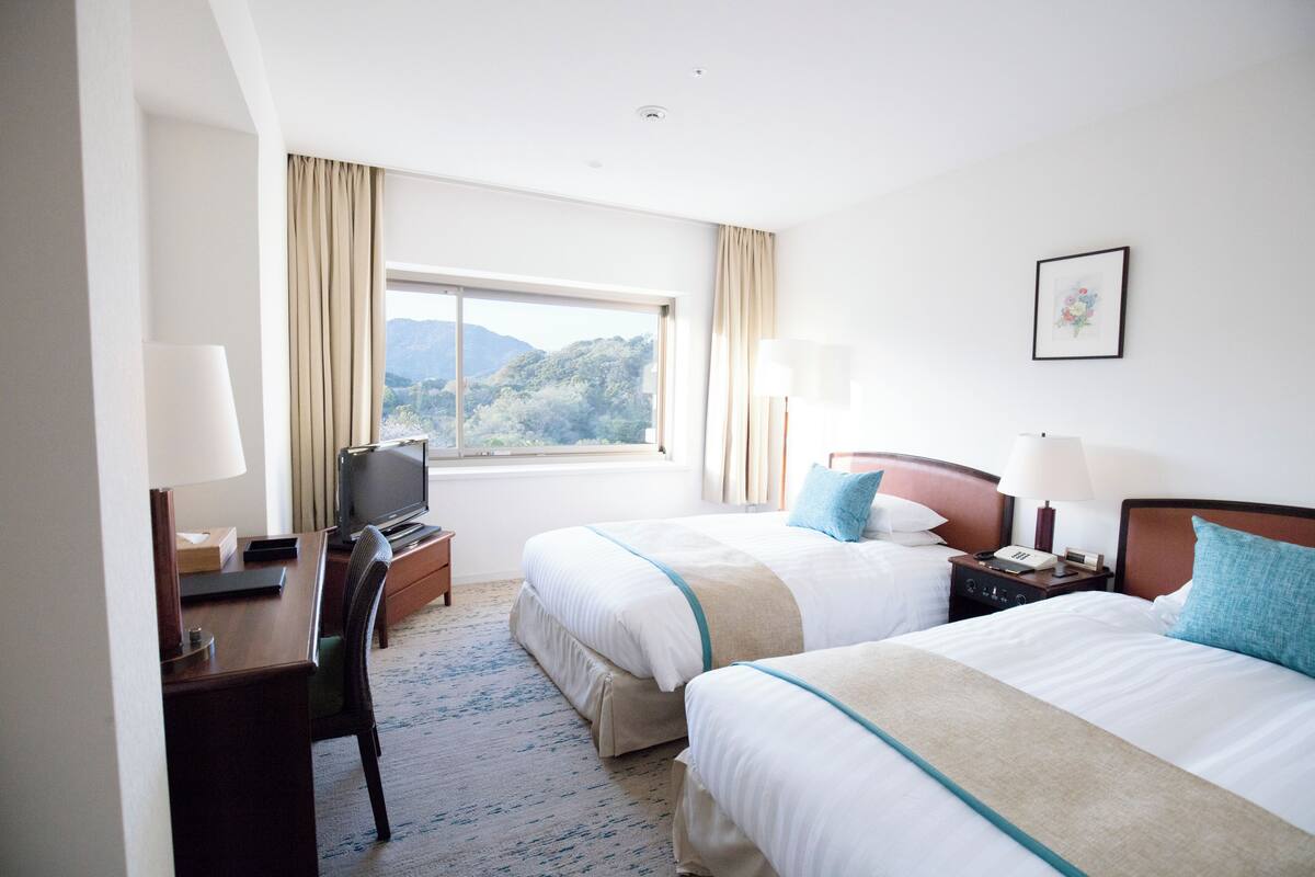 A hotel room with two beds, a desk, a TV, and a window with a mountain view.