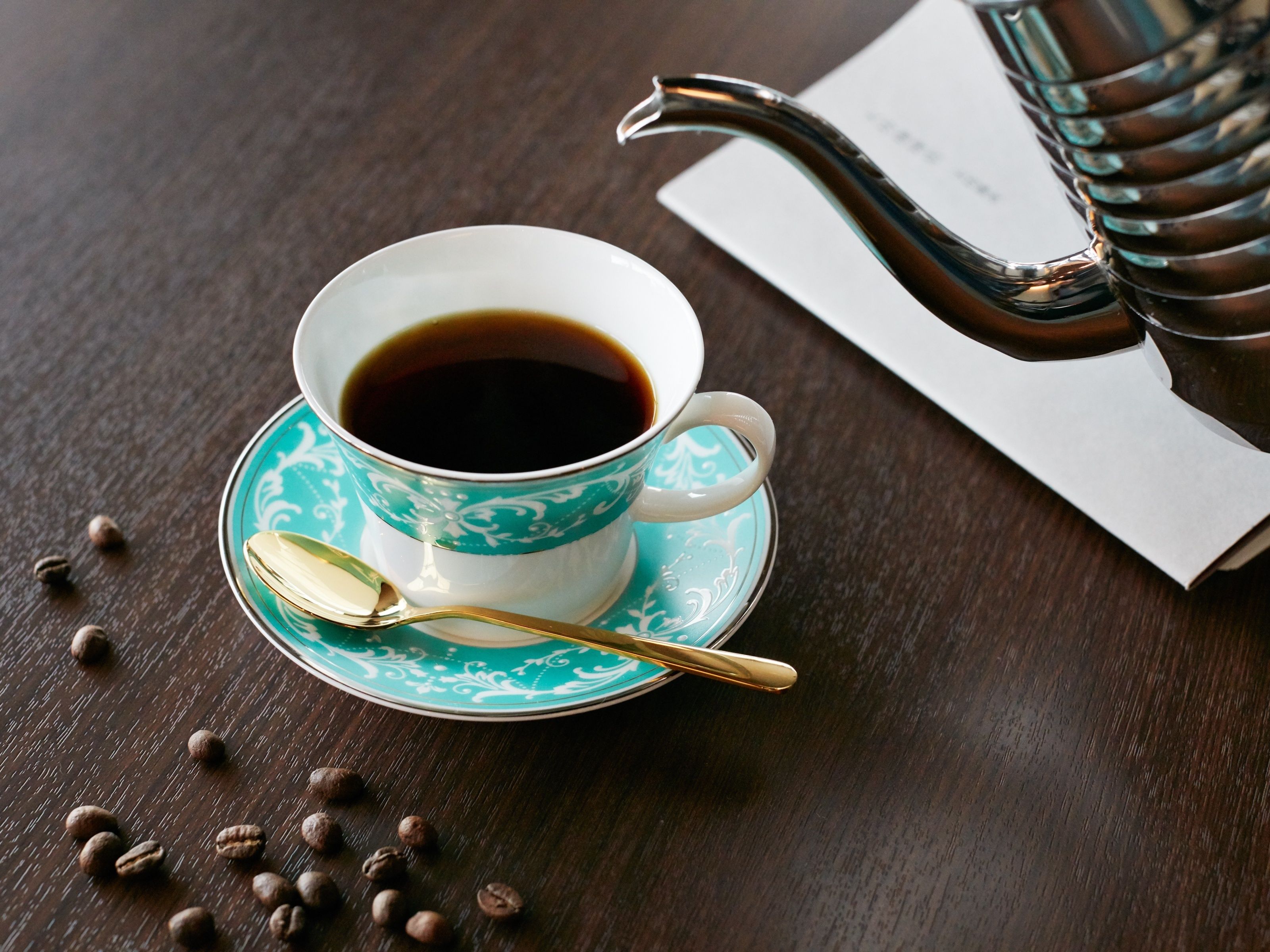 A cup of coffee on a saucer with a spoon, surrounded by coffee beans on a wooden table.
