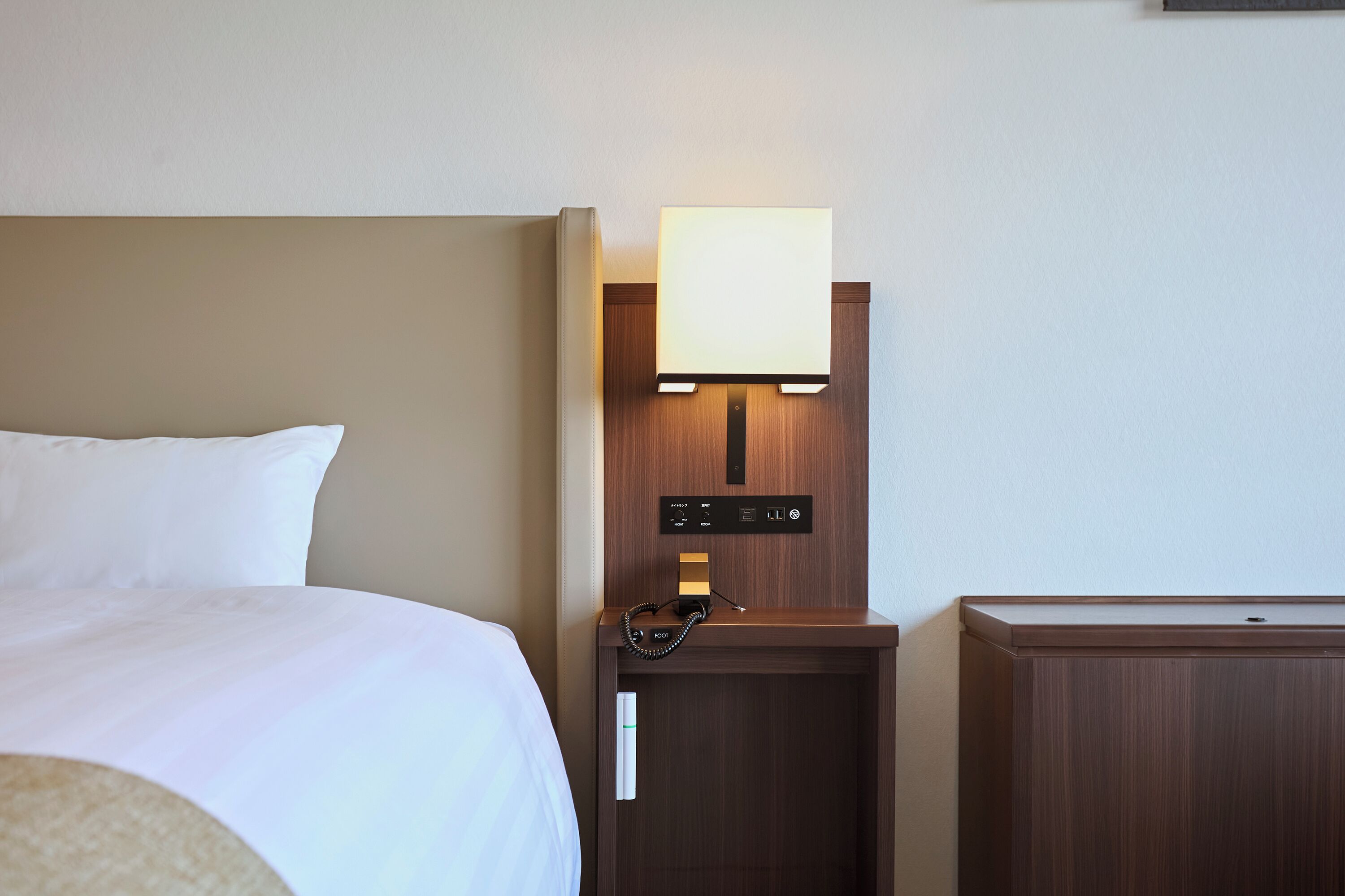 Una camera d'albergo con un letto, un comodino con una lampada, un telefono e una piccola credenza.
