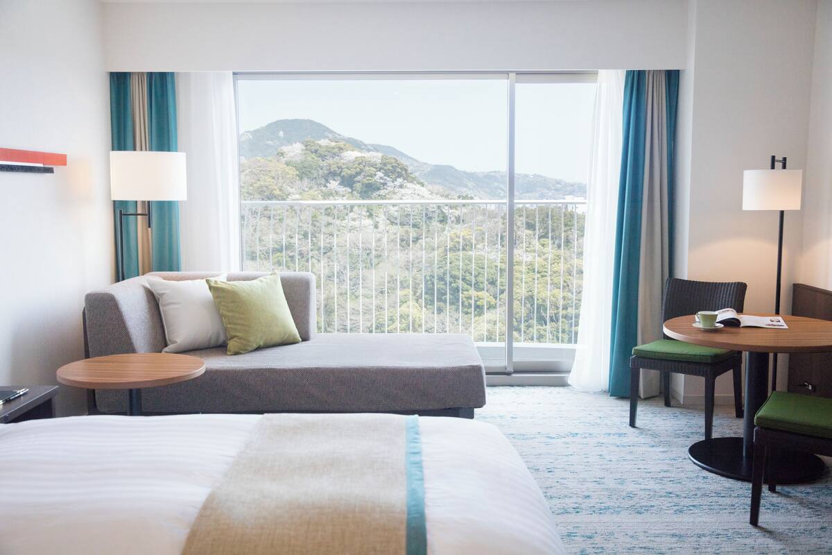 A hotel room with a bed, a sofa, a small table, and a chair. There is a large window with a view of mountains.