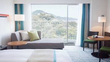 Habitación de hotel con cama, sofá, una mesita y una silla. Amplia ventana con vistas a las montañas.
