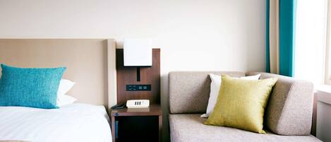 A hotel room with a bed, a nightstand with a phone and a clock, and a sofa with pillows.
