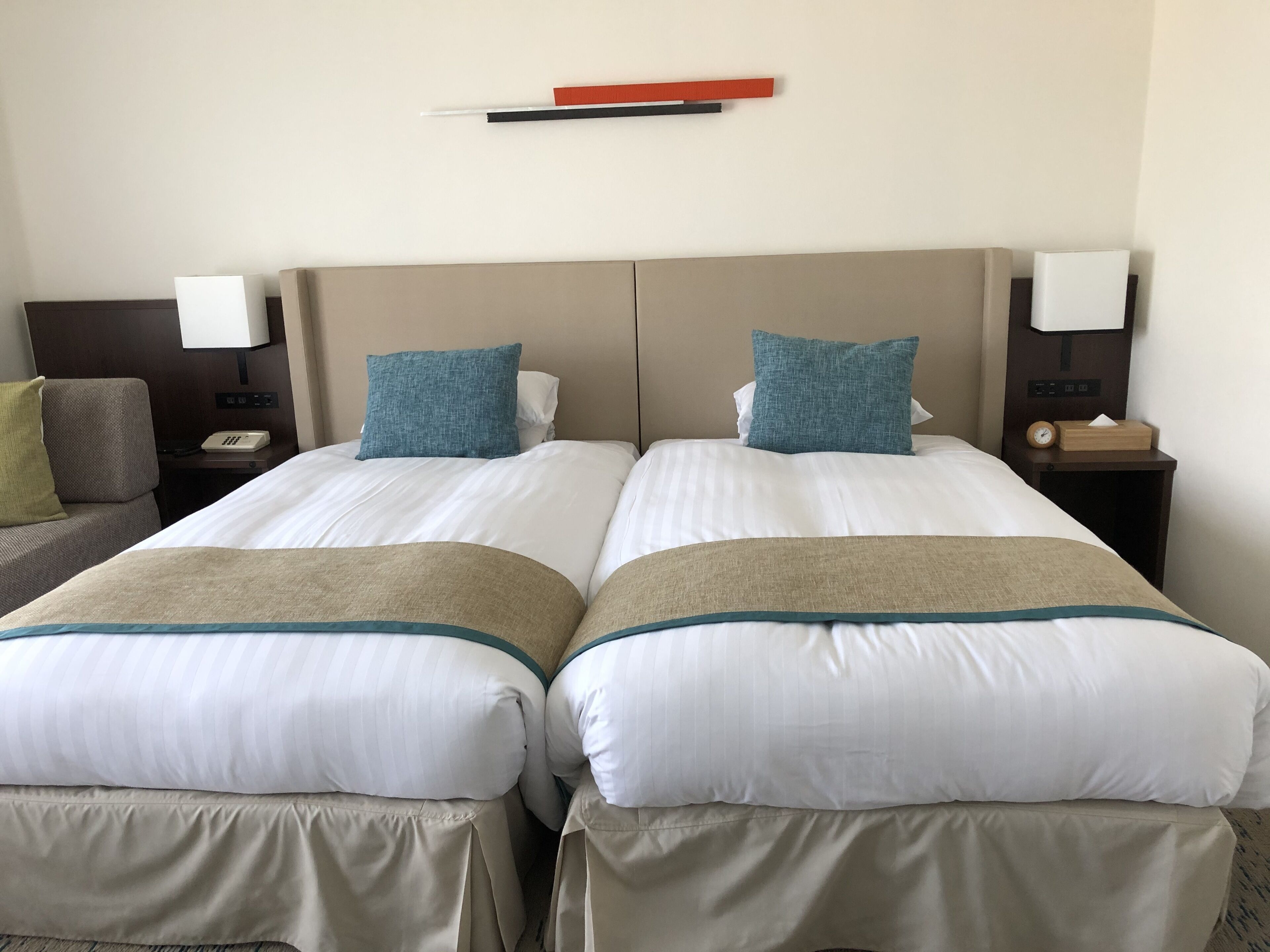 A hotel room with two beds, each with a white and beige bedspread, blue pillows, and a wooden headboard. There is a nightstand with a lamp and a clock on the right side.
