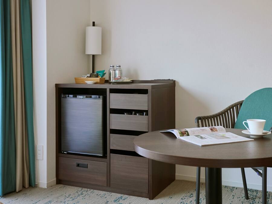 A compact hotel room with a bed, a bedside table with a lamp, a small cabinet, and a round table with a chair.