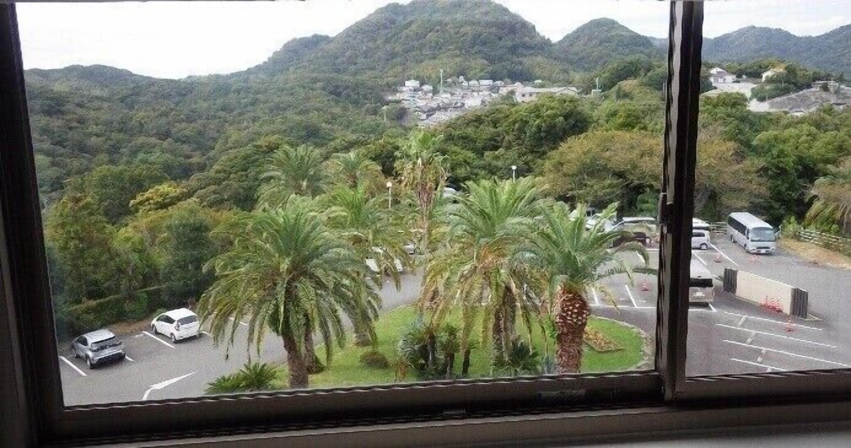 Vista desde una ventana sobre un estacionamiento con palmeras y un paisaje montañoso al fondo.