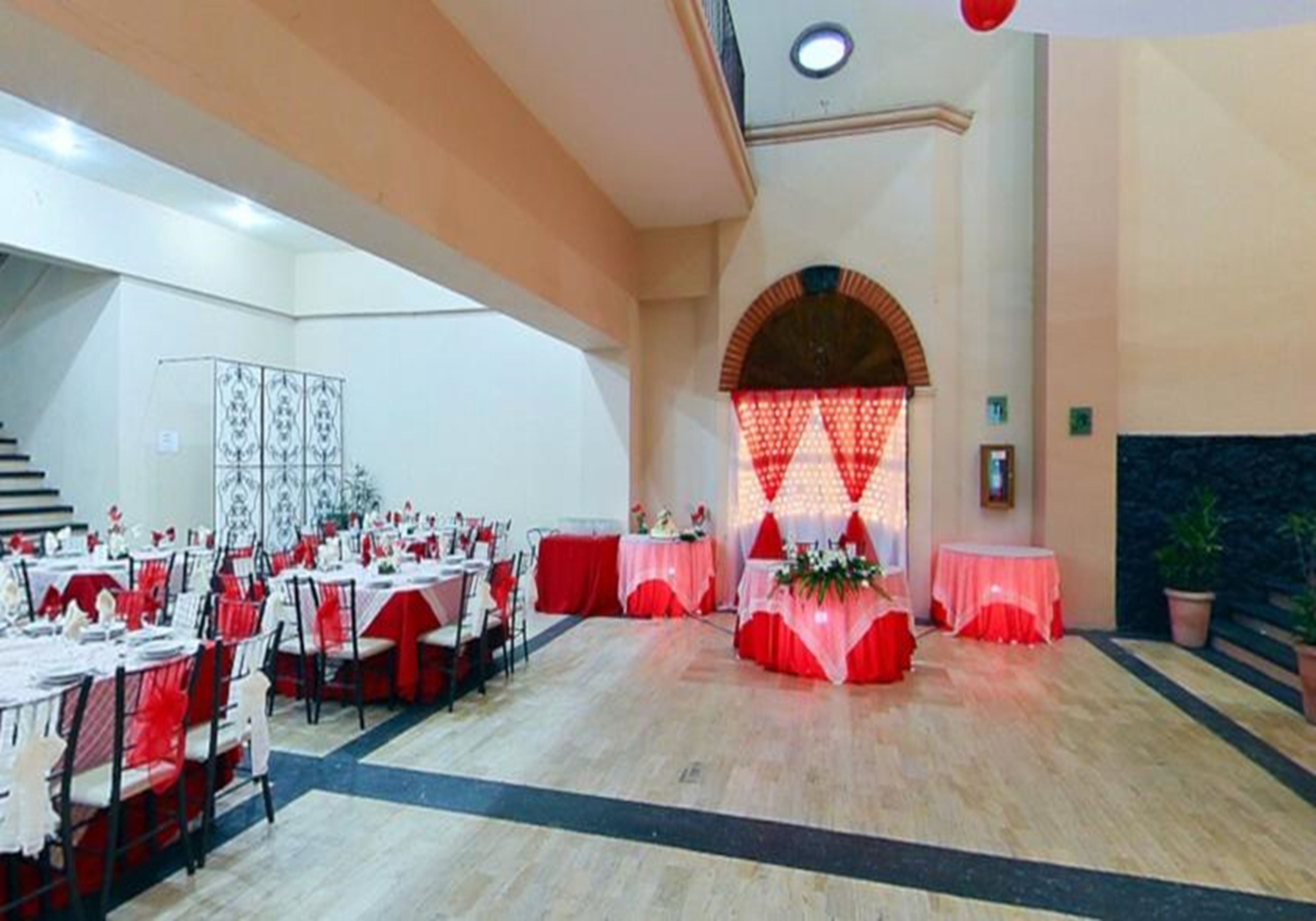 Un salón de banquetes con varias mesas redondas, manteles blancos y rojos, y un escenario con un arreglo floral.