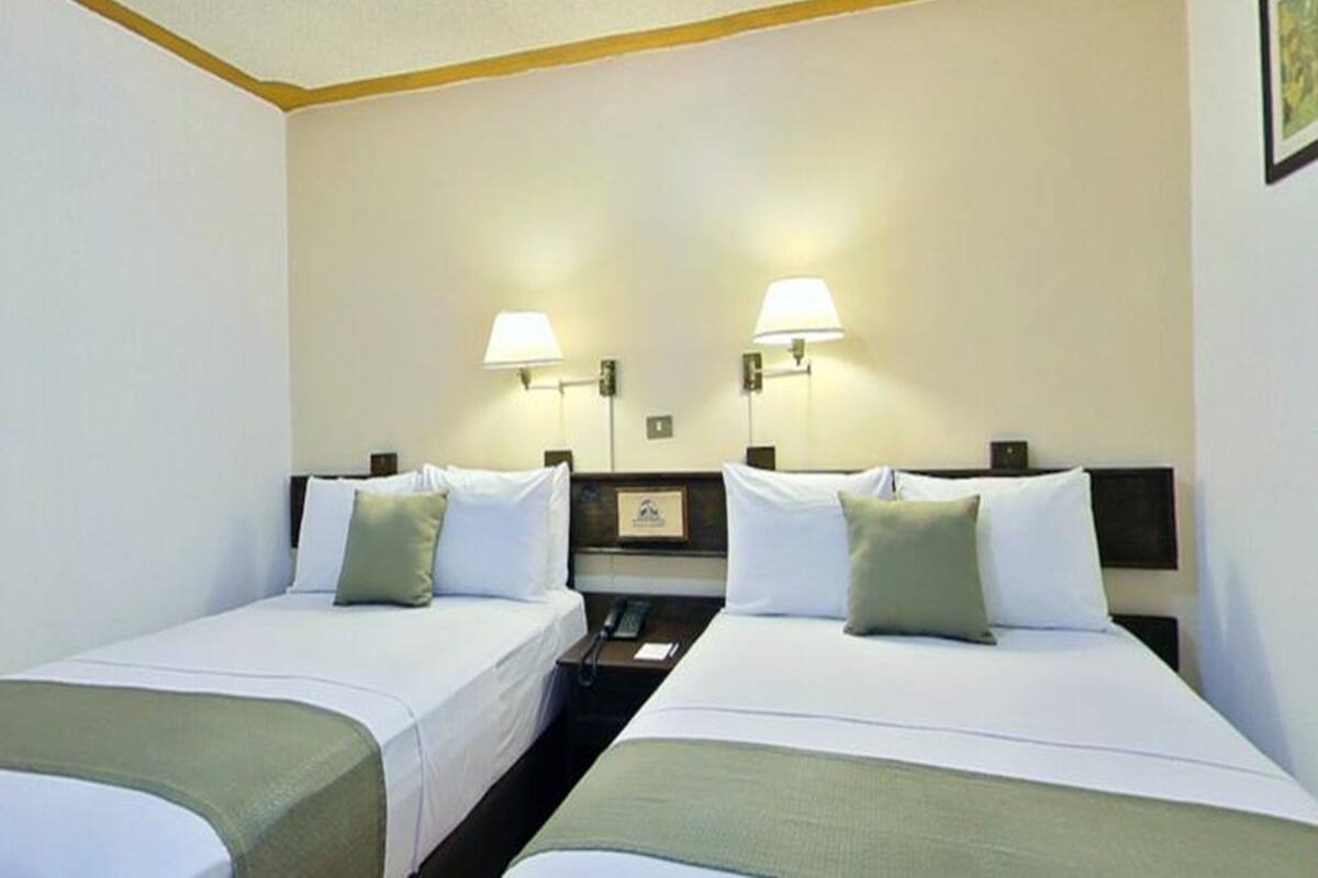 A hotel room with two single beds, each with a pillow and a blanket. There are two wall-mounted lamps on either side of the beds.