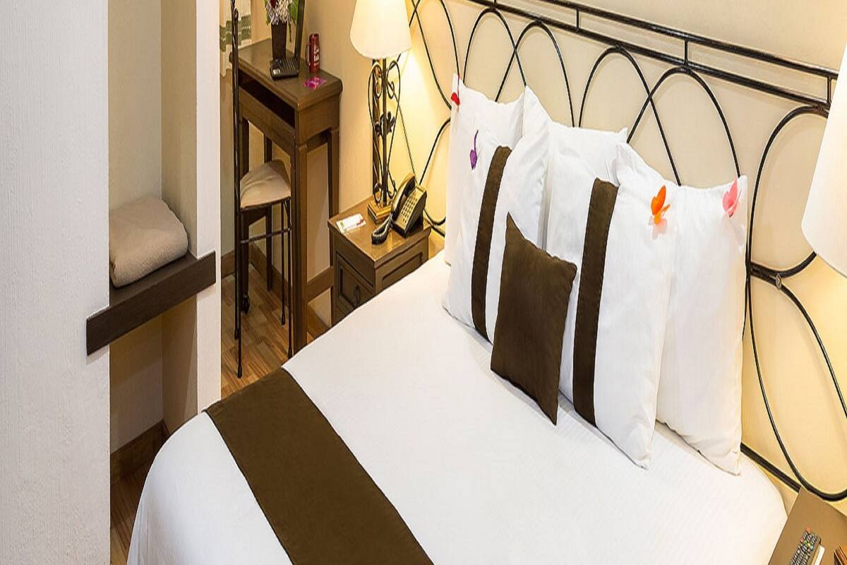 A hotel room with a bed, bedside table, lamp, chair, and a desk with a telephone and a vase of flowers.