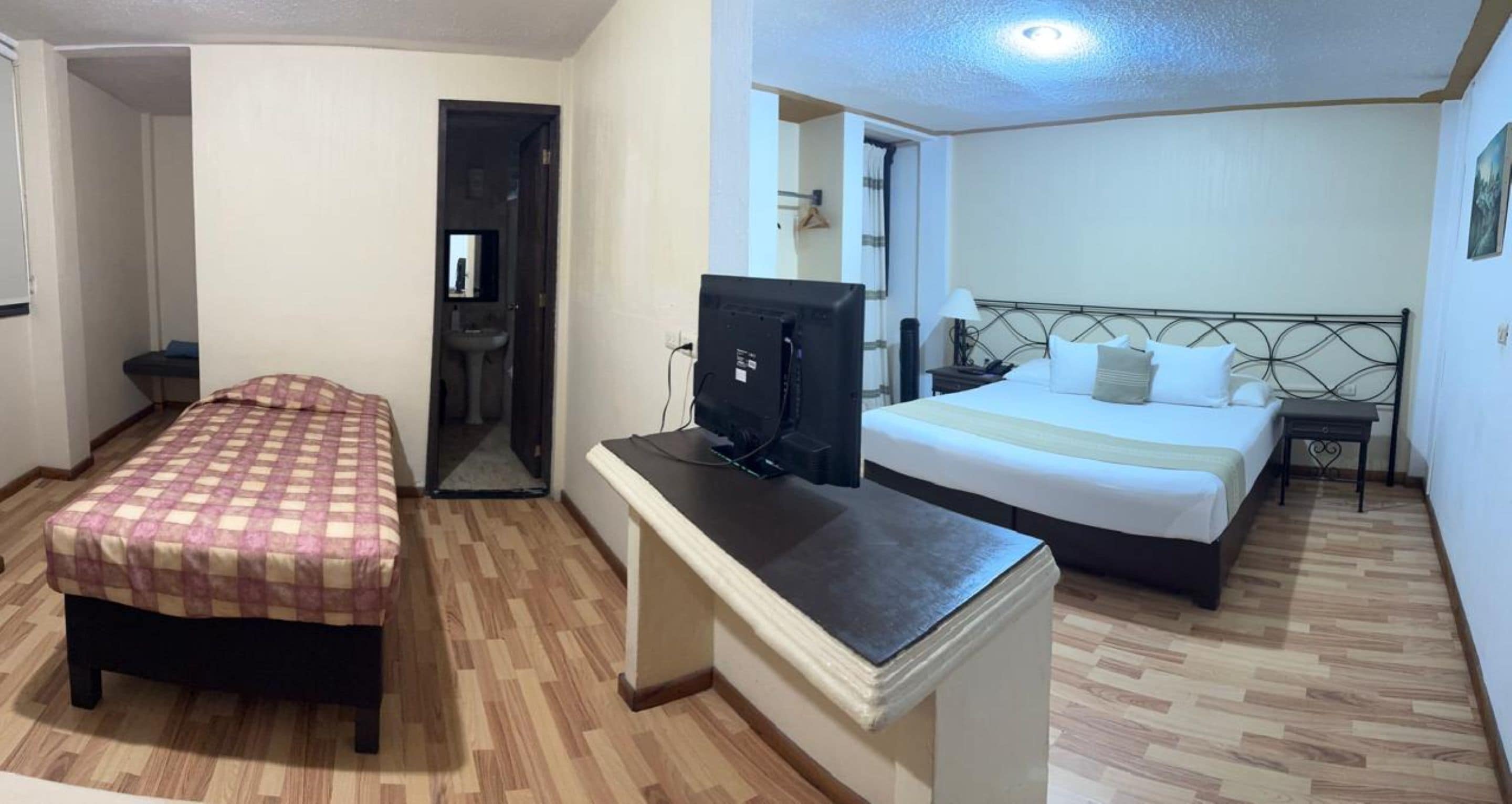 Quarto de hotel com uma cama, uma escrivaninha com computador, uma mesinha e um banheiro visível através de uma porta aberta.