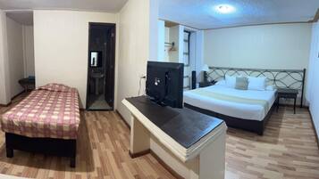 Quarto de hotel com uma cama, uma escrivaninha com computador, uma mesinha e um banheiro visível através de uma porta aberta.