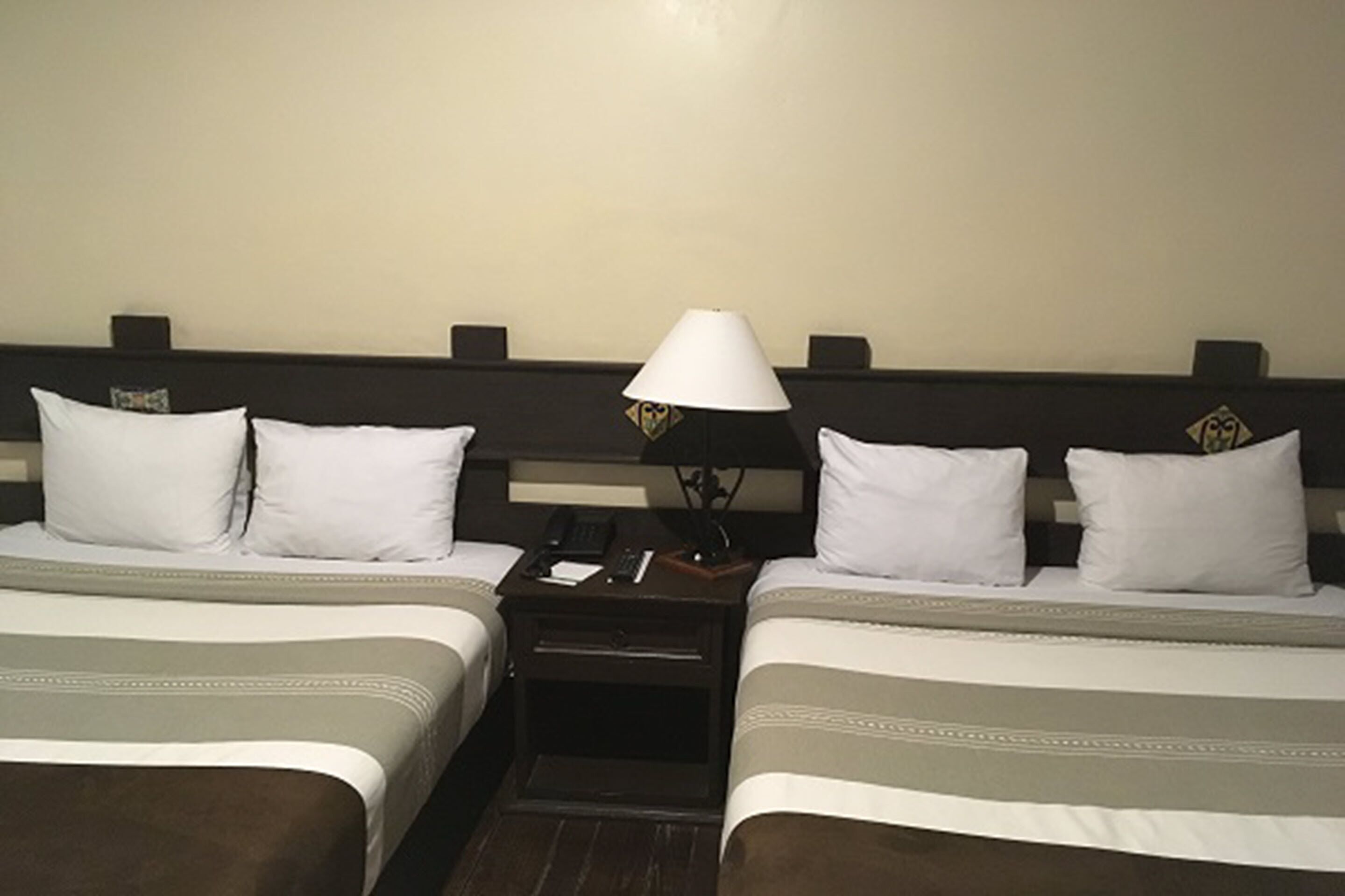 A hotel room with two beds, each with white bedding and a brown headboard. There is a nightstand between the beds with a lamp and a phone.
