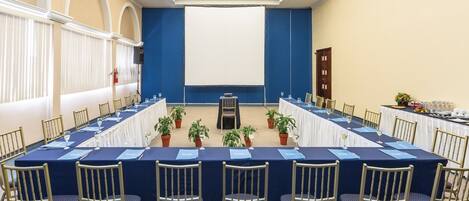 Un salón de conferencias con una larga mesa, sillas, plantas en macetas y una pantalla de proyector.