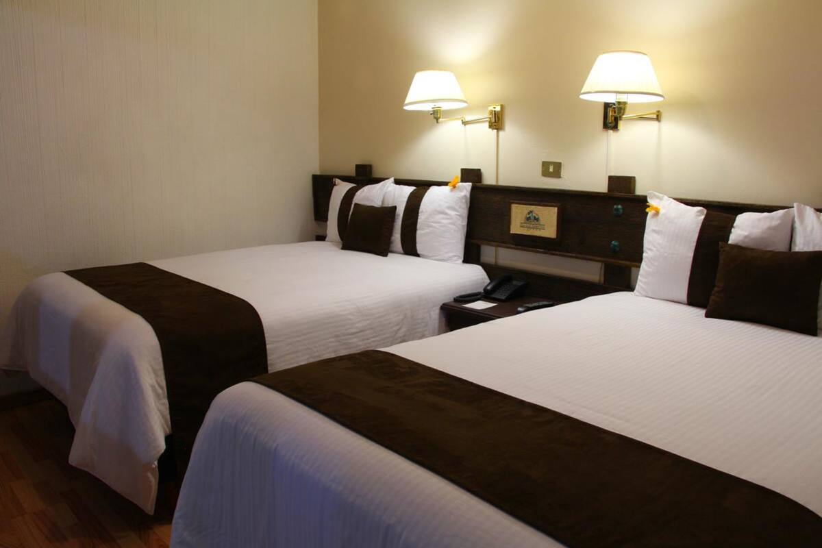 A hotel room with two beds, each with white bedding and brown headboards. There are two wall-mounted lamps on either side of the beds.