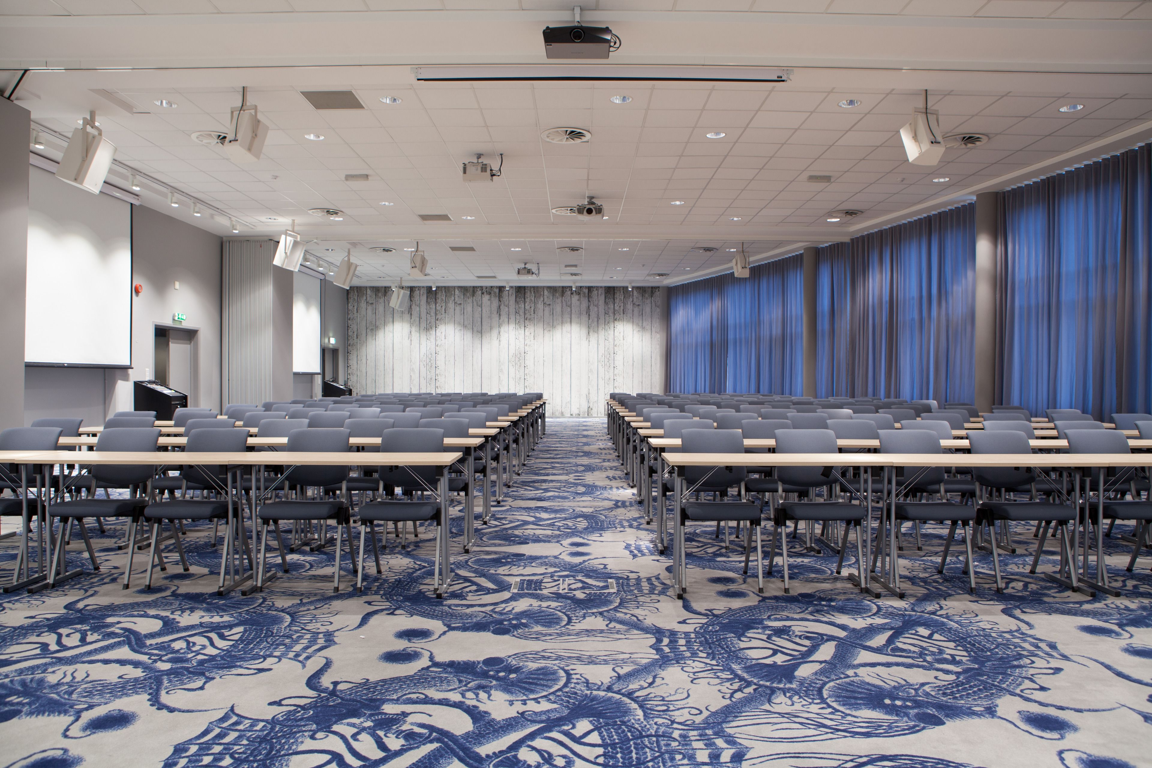 Un salón de conferencias con filas de mesas y sillas, una alfombra estampada y una pantalla de proyector.