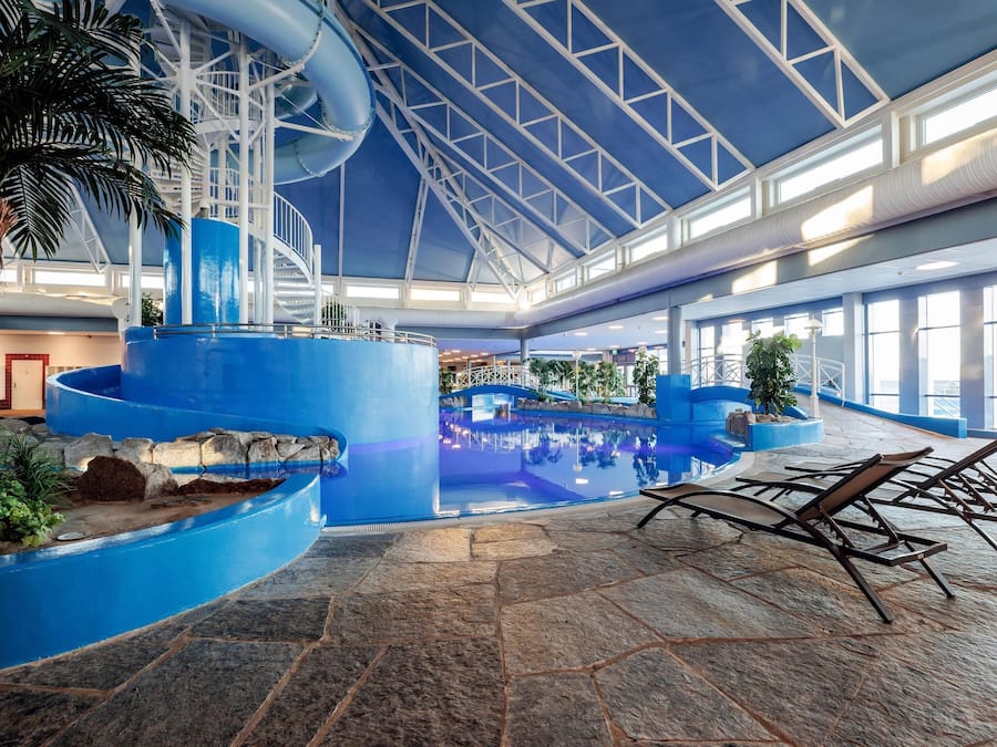An indoor swimming pool with a blue color scheme, a palm tree, lounge chairs, and a high ceiling with skylights.