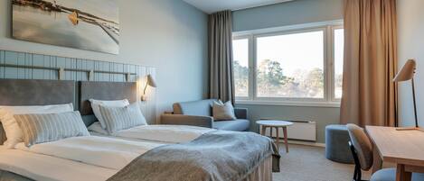 Quarto de hotel com cama, sofá, mesa pequena e cadeira. Janela com cortinas e vista para árvores.